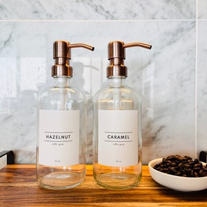 May include: Two clear glass syrup dispensers with copper-colored pumps labeled "Hazelnut" and "Caramel". The dispensers sit on a wooden tray with a small white bowl of coffee beans. The background is a marble tile wall.