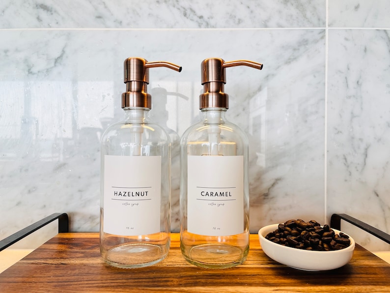 May include: Two clear glass syrup dispensers with copper-colored pumps labeled "Hazelnut" and "Caramel". The dispensers sit on a wooden tray with a small white bowl of coffee beans. The background is a marble tile wall.