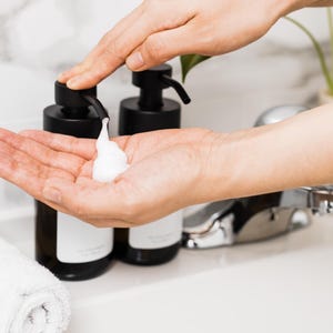 May include: A person's hand pumps a black pump bottle of liquid soap into their other hand. The soap is white and foamy. There are two black pump bottles of soap on the counter.