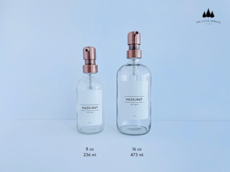 May include: Two clear glass bottles with copper-colored pump tops. Each bottle has a white label that reads "HAZELNUT coffee syrup." The smaller bottle is labeled 8 oz (236 ml), and the larger bottle is labeled 16 oz (473 ml).