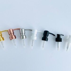 May include: An assortment of soap and lotion dispenser pumps in various colors, including gold, copper, silver, white, and black. Each pump has a clear plastic tube and a curved spout. The pumps are arranged in a row against a white background.