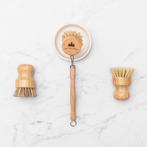 May include: Three wooden dish brushes with natural bristles. The brush on the left has a round head, the brush in the middle has a long handle and a round head, and the brush on the right has a round head. The brush in the middle has the text "The Little Spruce" engraved on the handle.