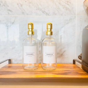May include: Two clear glass syrup bottles with gold pump tops labeled "Caramel" and "Vanilla" on a wooden tray. A coffee grinder is visible on the right. The bottles are for coffee syrup.