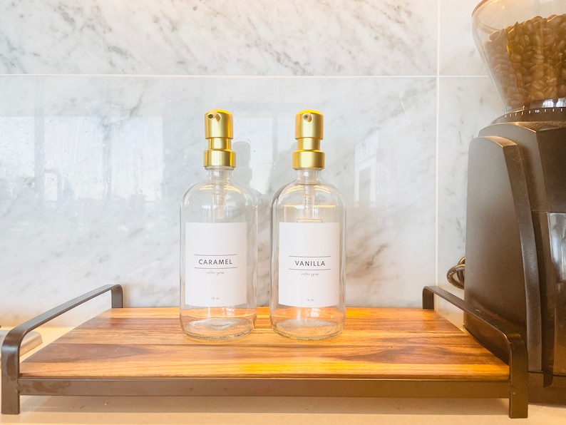 May include: Two clear glass syrup bottles with gold pump tops labeled "Caramel" and "Vanilla" on a wooden tray. A coffee grinder is visible on the right. The bottles are for coffee syrup.
