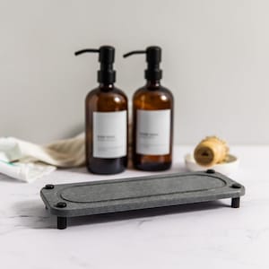 Peut inclure: Un porte-savon rectangulaire gris avec des bords arrondis et quatre pieds noirs. Le porte-savon est fait d'un matériau texturé et a un ovale surélevé au centre. Deux flacons en verre ambré avec des pompes noires sont en arrière-plan.