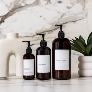 May include: A set of brown pump bottles labelled "Shampoo", "Body Wash", and "Conditioner" with white labels. A white decorative vase and a small potted plant are also present. The bottles are arranged on a marble surface.