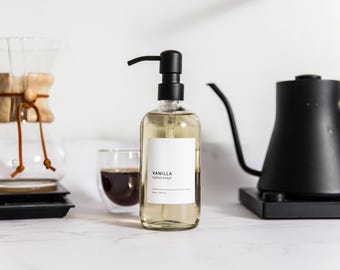 May include: A clear glass bottle with a black pump dispenser filled with vanilla coffee syrup. The label reads "Vanilla Coffee Syrup".