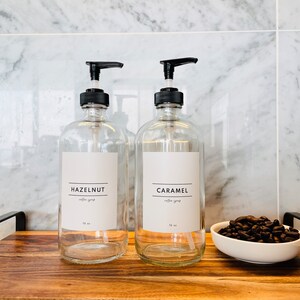 May include: Two clear glass syrup dispensers with black pump tops, labeled "HAZELNUT" and "CARAMEL". A small white bowl of coffee beans sits on a wooden tray. The dispensers are 16 oz.