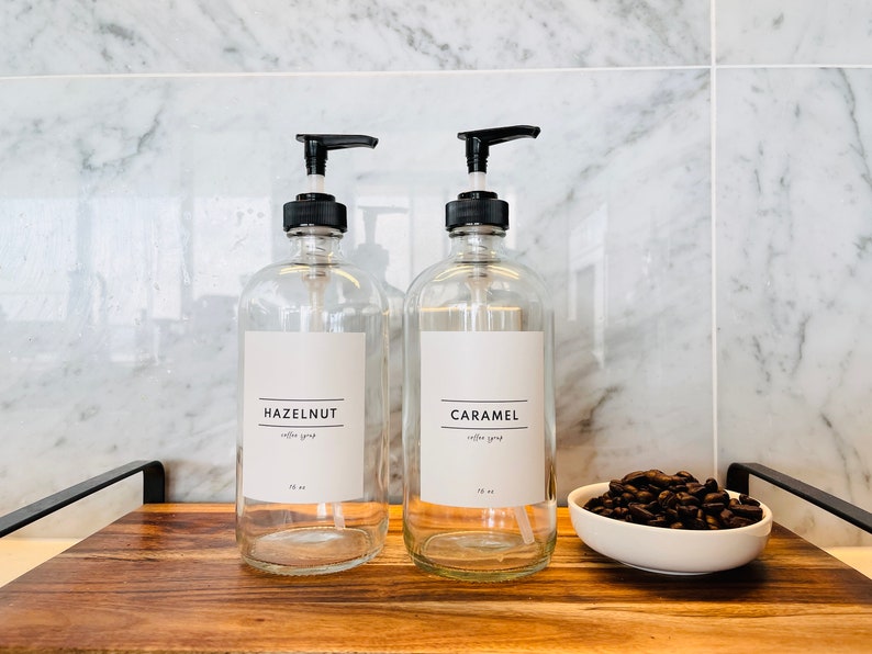 May include: Two clear glass syrup dispensers with black pump tops, labeled "HAZELNUT" and "CARAMEL". A small white bowl of coffee beans sits on a wooden tray. The dispensers are 16 oz.