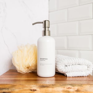 May include: White ceramic shampoo bottle with a silver pump dispenser on a wooden surface. The bottle has the word "SHAMPOO" printed on it. There is a yellow bath sponge and a white towel next to the bottle.