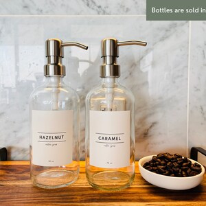 May include: Two clear glass bottles with silver pump dispensers labeled "HAZELNUT" and "CARAMEL coffee syrup". Each bottle is 16 oz. A small white bowl filled with coffee beans sits on a wooden tray. The text "Bottles are sold individually" is visible.