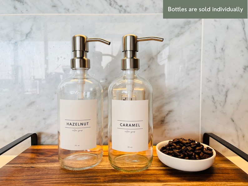 May include: Two clear glass bottles with silver pump dispensers labeled "HAZELNUT" and "CARAMEL coffee syrup". Each bottle is 16 oz. A small white bowl filled with coffee beans sits on a wooden tray. The text "Bottles are sold individually" is visible.