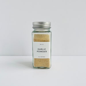 以下が含まれることがあります： 銀色の蓋が付いた透明なガラス瓶で、薄茶色のガーリックパウダーが入っています。前面の白いラベルには「GARLIC POWDER」と「ALL NATURAL」と書かれています。瓶は長方形で、白い表面に置かれています。
