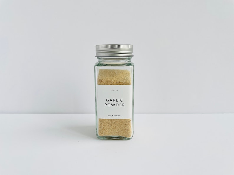以下が含まれることがあります： 銀色の蓋が付いた透明なガラス瓶で、薄茶色のガーリックパウダーが入っています。前面の白いラベルには「GARLIC POWDER」と「ALL NATURAL」と書かれています。瓶は長方形で、白い表面に置かれています。