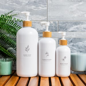 May include: Three white pump bottles with bamboo lids labelled "Hand Soap", "Conditioner", and "Body Wash". Each bottle has a minimalist floral design. The bottles are on a wooden surface with a green cactus and fern.