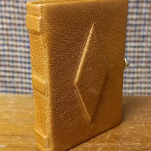 Puede incluir: Un pequeño libro de cuero marrón claro con una hendidura en forma de diamante en la portada. El libro está de pie sobre una superficie de madera, con una tela estampada en el fondo. El libro tiene un cierre en el lateral.