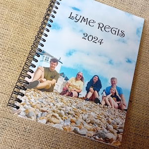 Può includere: Quaderno a spirale con una foto di gruppo su una spiaggia. La copertina mostra la scritta "LYME REGIS 2024" su sfondo cielo. Il quaderno è aperto, rivelando l'immagine delle persone sedute sulla spiaggia.