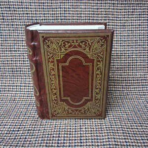 May include: A hardcover book with a burgundy cover and gold filigree design. The spine is also burgundy with gold accents. The book is closed and standing upright, showcasing the ornate cover details. The pages are white.