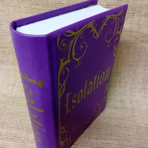 May include: A purple book with the word "Isolation" in gold lettering. The book has gold decorative flourishes around the edges. The pages are white, and the cover is a solid, deep purple.