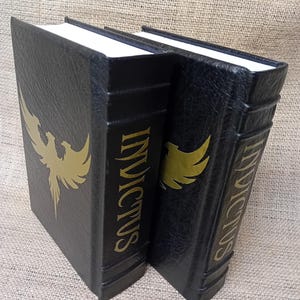 May include: Two black bookends with gold lettering and a gold phoenix design. The word "INVICTUS" is printed vertically on the spine. The bookends have white pages and are made of a textured material.