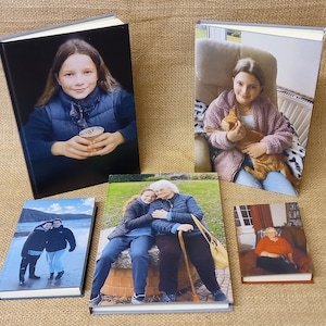 May include: A collection of hardcover photo books with various images. The books feature images of people, including a young girl holding a cup, a girl with a cat, and a group of people outdoors. The books are displayed on a textured, tan surface.