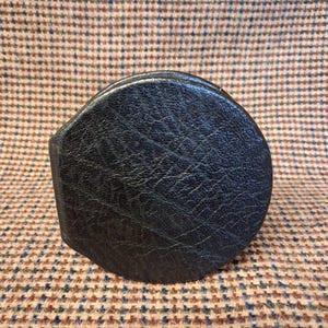 May include: A black leather coaster with a textured surface. The coaster has a rounded shape with a small, rectangular tab on one side. The background is a brown and blue patterned fabric.