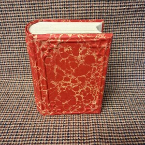 May include: A hardcover book with a red cover featuring a gold marbled design. The book is standing upright, showcasing its textured cover and the white pages. The book's spine is rounded, and the edges are gilded.