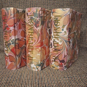 May include: Three hardcover books with marbled covers in shades of orange, pink, cream, and dark brown. The spines feature gold foil lettering that reads Ironheart I, Ironheart II, and Ironheart III, arranged in a vertical orientation on each book.