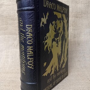 May include: A dark blue book with gold lettering and illustrations. The title "DRACO MALFOY" is prominently displayed, along with "and the mortifying ORDEAL OF BEING IN LOVE." The spine also features the title. The book has a textured cover.