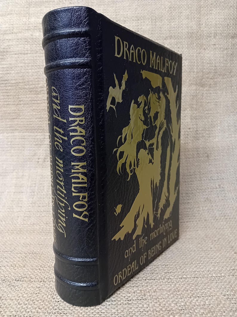 May include: A dark blue book with gold lettering and illustrations. The title "DRACO MALFOY" is prominently displayed, along with "and the mortifying ORDEAL OF BEING IN LOVE." The spine also features the title. The book has a textured cover.