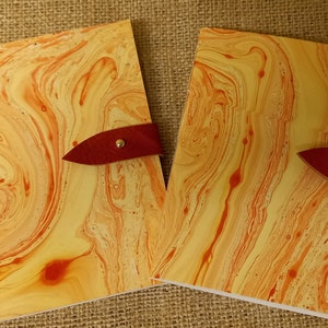 May include: Two yellow and orange marbled notebooks with red leather closures and gold metal accents. The notebooks are open and lying on a burlap surface.