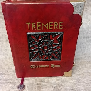 May include: A red leather-bound book featuring the word TREMERE in gold lettering above a black, intricate, geometric metal inlay. The name Theodore Hum is printed below in gold. The book has silver-toned metal corner protectors and a bookmark.