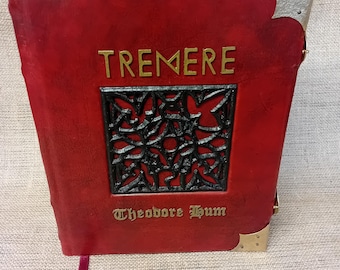 Pode incluir: Um livro encadernado em couro vermelho com a palavra TREMERE em letras douradas sobre uma incrustação metálica preta com design geométrico complexo. O nome Theodore Hum está impresso abaixo em dourado. Cantos metálicos prateados e um marcador.