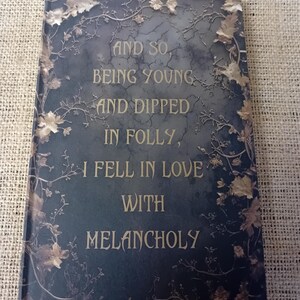 Pu&ograve; includere: Un diario grigio scuro con un design in rilievo dorato di foglie e viti. Il testo "AND SO BEING YOUNG AND DIPPED IN FOLLY, I FELL IN LOVE WITH MELANCHOLY" &egrave; stampato in oro sulla copertina.