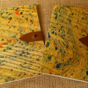 May include: Two yellow, green, and blue marbled notebooks with brown leather straps and brass accents. The notebooks are closed and lying on a brown burlap surface.