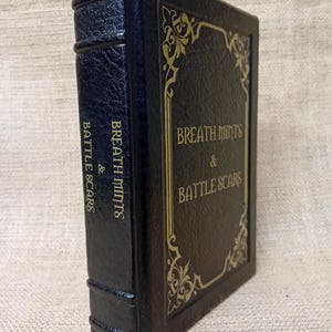 May include: A dark blue book with gold lettering and decorative accents. The book's cover reads "Breath Mints & Battle Scars" in an elegant font. The spine also features the title. The book has a textured, leather-like appearance.