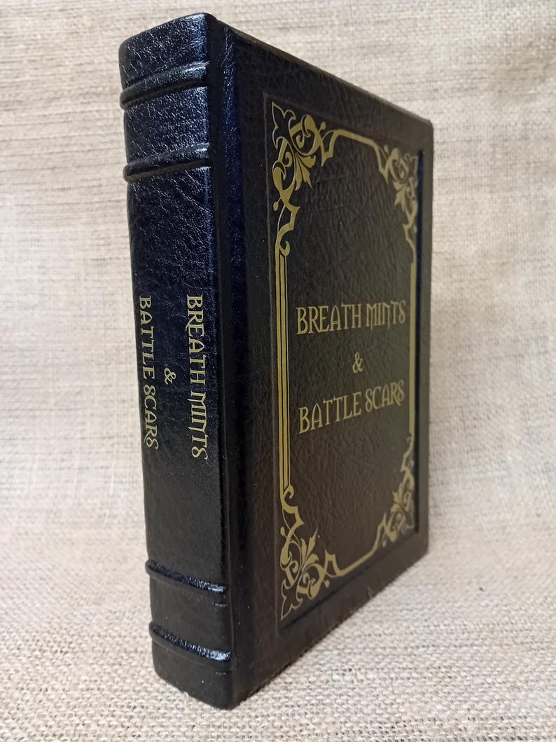 May include: A dark blue book with gold lettering and decorative accents. The book's cover reads "Breath Mints & Battle Scars" in an elegant font. The spine also features the title. The book has a textured, leather-like appearance.