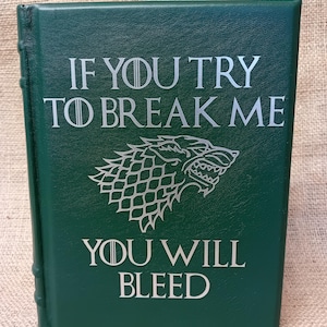 May include: A dark green book with silver lettering that reads "IF YOU TRY TO BREAK ME YOU WILL BLEED." A wolf emblem is centered below the text. The book has a textured cover and a spine with visible binding.
