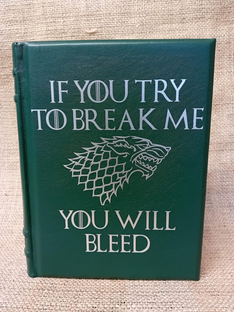 May include: A dark green book with silver lettering that reads "IF YOU TRY TO BREAK ME YOU WILL BLEED." A wolf emblem is centered below the text. The book has a textured cover and a spine with visible binding.