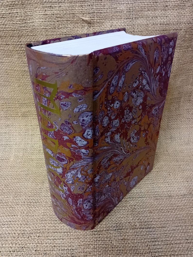 May include: A hardcover book with a marbled cover in shades of burgundy, gold, and white. The spine features gold lettering. The book's edges are gilded, and the pages are white.