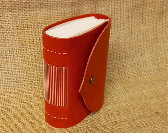 May include: A small red leather-bound journal with a flap closure and white stitching. The journal is closed and standing upright on a burlap surface.