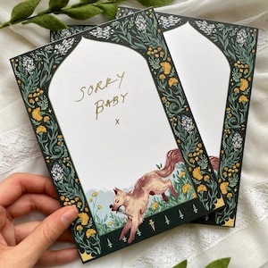 May include: Two greeting cards with a floral border in dark green, yellow, and white. The cards have the words "Sorry Baby" in gold script, and a drawing of a wolf with a knife. The cards are held by a hand.