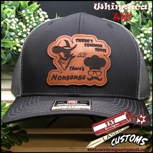 May include: A black trucker hat with a gray mesh back and a leather patch. The patch features a cowboy and a chef with the text "There's Common Sense And There's Nonsense". The hat is adjustable.