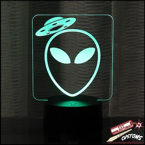May include: A glowing green alien head and UFO design night light. The alien has large almond-shaped eyes and is topped with a flying saucer. The light sits on a black base and is illuminated with a green glow.