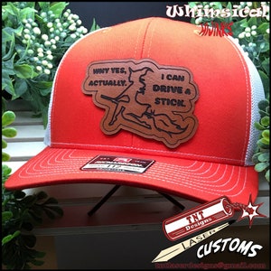 May include: An orange and white trucker hat with a leather patch. The patch features a witch riding a broom and the text "WHY YES, ACTUALLY. I CAN DRIVE A STICK." The hat has a curved brim and an adjustable snap closure.