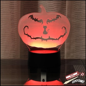 May include: A Halloween-themed LED lamp shaped like a jack-o'-lantern. The pumpkin has a carved face with bat and cat silhouettes. The lamp emits a red glow and sits on a black base. The text "TNT Designs Laser Customs" is visible.