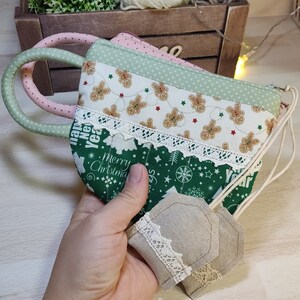 May include: A set of three green and white fabric pouches with a Christmas theme. The pouches have a gingerbread man pattern, a green and white Christmas tree pattern, and a beige fabric with lace trim. The pouches are held in a hand.