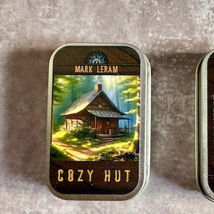 May include: Two rectangular metal tins. One tin features a cabin illustration with the text "COZY HUT" and "MARK LERAM". The other tin has text about the game, including a quote by Henry David Thoreau.