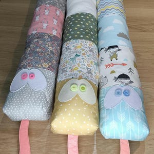 May include: Three colourful patchwork fabric baby toys shaped like snakes with button eyes and pink ribbon tails. The snakes are made of different patterned fabrics, including floral, polka dot, and geometric designs.