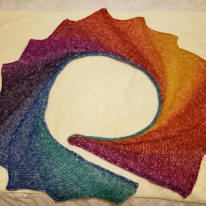 Puede incluir: Una bufanda de punto de colores arcoíris con un acabado brillante. La bufanda está doblada en forma de círculo y está sobre una superficie blanca.